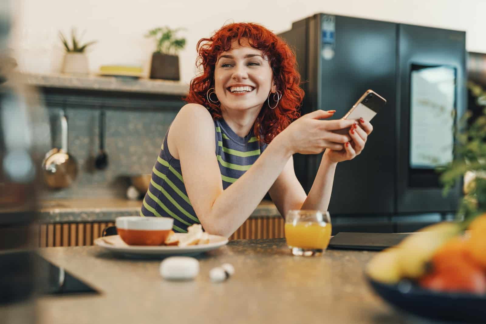Glad kvinna vid köksbordet som tittar på sin mobiltelefon